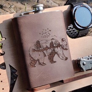 May include: A brown leather flask with a bear and forest design, a black smartwatch, and a folding knife rest on a wooden tray. The tray has cutouts of a hat, sunglasses, a mustache, and a bow tie.