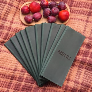 Puede incluir: Un conjunto de cubiertas de menú verde oscuro con la palabra "MENU" impresa. Una bandeja de madera contiene ciruelas y melocotones rojos. Los artículos están dispuestos sobre una manta estampada marrón rojizo.