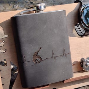 Puede incluir: Una petaca gris oscuro con tapa plateada, con un diseño de snowboarder y una línea de latidos del corazón. La petaca está sobre una superficie de madera con otros elementos decorativos. Mide aproximadamente 12 cm de alto.