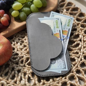 Puede incluir: Una cartera de cuero gris con un diseño festoneado único, que contiene varios billetes de 100 $. La cartera descansa sobre un mantel individual tejido, junto a una tabla de madera con uvas rojas y verdes, y una manzana roja.