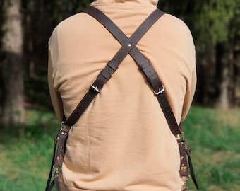 May include: A person wearing a brown leather camera strap with two camera bags attached. One bag is a cylindrical shape and the other is a rectangular shape. The person is wearing a tan hoodie and black trousers.