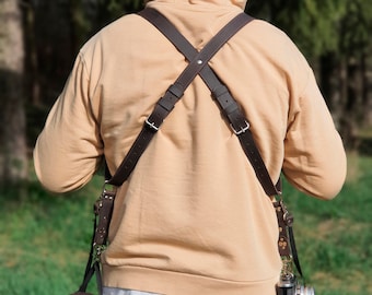 May include: A person wearing a brown leather camera strap with a camera and a brown leather pouch attached to it. The strap is worn over the shoulders and crosses at the back.