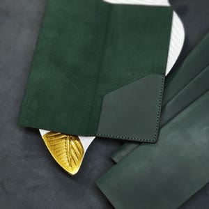Puede incluir: Un juego de cuatro carteras de cuero verde, una con una solapa de gamuza. Las carteras están dispuestas sobre una superficie gris oscuro.