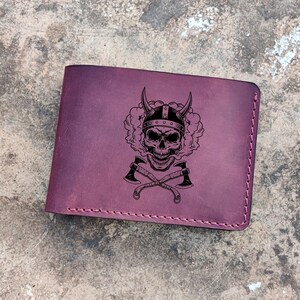 Puede incluir: Una cartera de cuero morado con una calavera con un casco con cuernos y hachas cruzadas grabadas en la parte delantera.