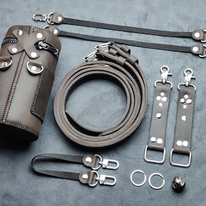 Può includere: Una borsa per fotocamera in pelle marrone con chiusura a zip e più cinturini. La borsa ha una forma cilindrica ed è decorata con accenti in metallo argentato. La borsa è mostrata su una superficie grigia testurizzata.