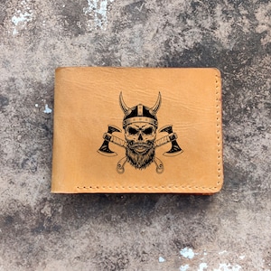 Puede incluir: Una cartera de cuero marrón con una ilustración en blanco y negro de una calavera con un casco con cuernos y hachas cruzadas.