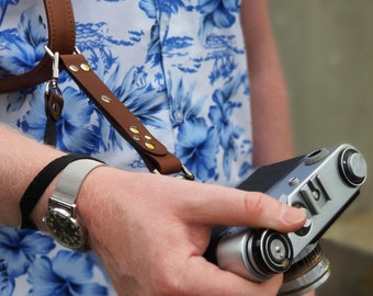 May include: A person wearing a white and blue Hawaiian shirt with a brown leather camera strap. The person is holding a silver camera with a black leather grip.
