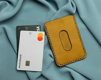 May include: A brown leather card holder with a stitched edge and a cut-out for a card. The card holder is on a blue fabric surface. Two credit cards are visible, one is black with the word "VISA" and the other is white with the word "monobank" and the Mastercard logo.