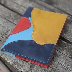 May include: A leather-bound journal with a patchwork cover featuring blue, red, and yellow panels. The panels are stitched together with a contrasting thread.