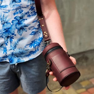 May include: A burgundy leather cylindrical bag with a black zip and a small front pocket. The bag has a matching burgundy leather strap with brass-coloured hardware. The person is wearing a blue and white floral shirt.