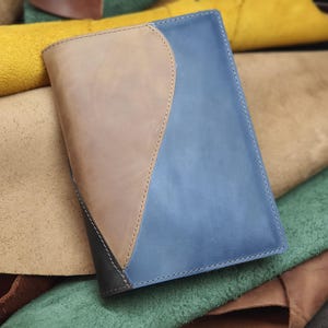 Może przedstawiać: Skórzany notes w kolorze niebieskim, brązowym i czarnym z pikowanym wzorem. Notes leży na stosie kawałków skóry w różnych kolorach.