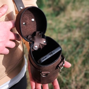 May include: A brown leather cylindrical phone case with a zipper closure and a strap. The case is open and a black smartphone is inside.