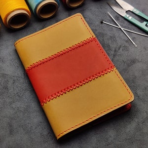 Könnte beinhalten: Ein braunes Ledernotizbuch mit einem roten Lederstreifen und dekorativen Nähten. Das Notizbuch liegt auf einer grauen Oberfläche.