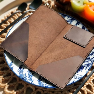 May include: Open brown leather wallet with card slots, on a decorative plate with blue and white floral patterns. The wallet is surrounded by a woven placemat, black utensils, and candles.