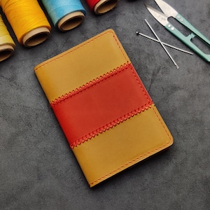 Könnte beinhalten: Ein gelbes Ledernotizbuch mit einem roten Lederpanel und dekorativen Nähten. Das Notizbuch liegt auf einer grauen Oberfläche mit Garnrollen und Nähwerkzeugen.