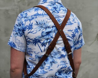 May include: Brown leather suspenders with adjustable buckles cross over the back of a person wearing a short-sleeved shirt. The shirt has a white background with a blue floral and palm tree pattern. The suspenders are attached with metal rivets.