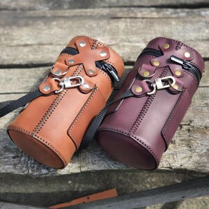 Peut inclure: Deux sacs en cuir cylindriques avec une fermeture à glissière et une sangle. Un sac est brun et l'autre est bordeaux.