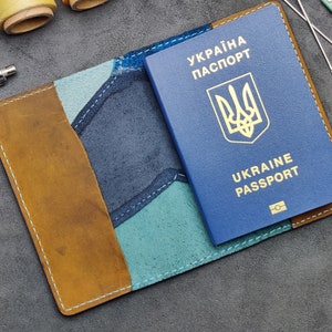 Könnte beinhalten: Ein braunes Leder-Reisepass-Etui mit blauen und grünen Akzenten. Das Etui hat ein genähtes Design und ist geöffnet, um einen blauen ukrainischen Reisepass mit dem Text "UKRAINE PASSPORT" und dem ukrainischen Wappen zu zeigen.