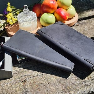 Peut inclure: Deux portefeuilles en cuir gris foncé sont présentés sur une surface en bois rustique. Les portefeuilles sont rectangulaires avec des bords cousus. Un petit pot en verre avec une bougie, une assiette en bois avec des pommes et des fleurs sont également dans l'image.