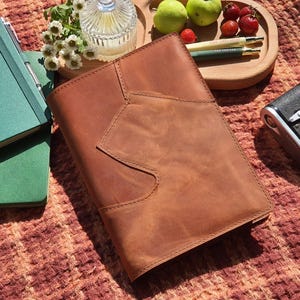 Può includere: Una copertina per diario in pelle marrone dal design unico, accanto a un taccuino verde e una fotocamera vintage. Un vassoio di legno contiene frutta, penne e un piccolo contenitore di vetro. Gli oggetti sono disposti su una superficie a motivi.
