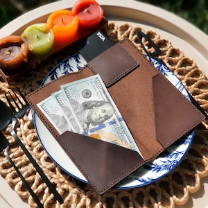 Op de afbeelding: Een bruine leren portemonnee met drie biljetten van 100 dollar in de voorzak. Een zwarte creditcard zit in de achterzak. De portemonnee ligt op een witte plaat met een blauw bloemenontwerp, met zwart bestek en kaarsen.
