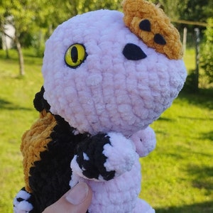 Può includere: Un giocattolo di peluche rosa e nero all'uncinetto con un occhio giallo e un cappello di uncinetto marrone. Il giocattolo è tenuto nella mano di una persona.