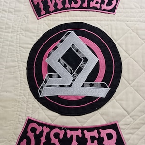 Op de afbeelding: Geborduurde patches met de woorden "TWISTED" en "SISTER" in roze letters op zwarte stof. Een cirkelvormige patch heeft een zilver en zwart geometrisch ontwerp met roze en zwarte concentrische cirkels. De patches liggen op een gewatteerde, gebroken witte achtergrond.