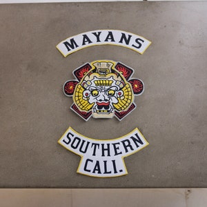 May include: Three embroidered patches with the words "MAYANS" and "SOUTHERN CALI." The central patch features a stylized face with yellow, red, and black details. The patches are on a neutral-toned background.