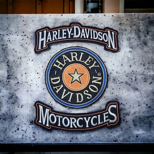 Peut inclure: Une surface grise texturée avec un logo Harley-Davidson en noir, blanc et orange. Le logo est entouré d'un cercle bleu avec les mots "Harley-Davidson" dans une arche noire au-dessus et "Motorcycles" dans une arche noire en dessous.