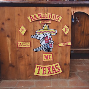 May include: A wooden sign with yellow and red patches. The patches have the words "Bandidos", "1%er", "2nd Bandido", "Texas", "MC", and "Texas". There is a cartoon image of a person wearing a sombrero and holding a sword.