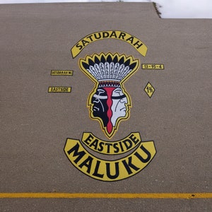 Pode incluir: Emblema pintado numa superfície castanha com as palavras "SATUDARAH" e "EASTSIDE MALUKU" em amarelo. O desenho central apresenta dois perfis nativos americanos, um preto e outro branco, com um cocar de penas.