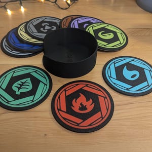 Op de afbeelding: Set van acht ronde onderzetters met verschillende gekleurde ontwerpen. Elke onderzetter heeft een zwarte achtergrond met een zeshoekige vorm in het midden. De ontwerpen omvatten een groen blad, een rode vlam, een blauwe vlam, een gele zon, een paarse bloem, een grijze driehoek, een bruine cirkel en een zwarte cirkel. De onderzetters zijn in een cirkel rond een zwarte cilindrische container gerangschikt.