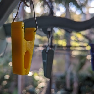 May include: A yellow plastic clothespin hanging from a wire clothesline. The clothespin is designed to hold clothes or other items securely.