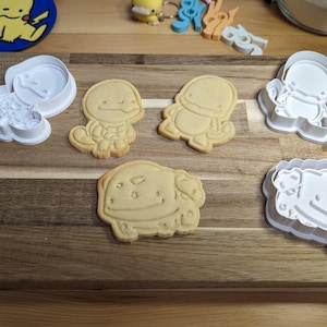 May include: A wooden cutting board displays several cookies and cookie cutters. The cookies are shaped like cartoon characters. White plastic cookie cutters of the same characters are also present. A Pikachu sticker is in the background.
