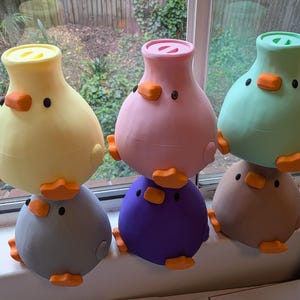 Puede incluir: Cinco recipientes redondeados con forma de pato, con picos y patas naranjas. La fila superior presenta figuras amarillas, rosas y verde claro, mientras que la inferior incluye una gris y una morada. Cada una tiene una tapa circular y ojos negros.