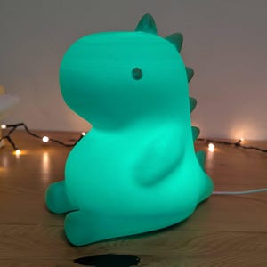 Può includere: Una luce notturna a forma di dinosauro verde acqua. La lampada ha un design liscio e arrotondato con un piccolo occhio e una fila di spine dorsali. È posizionata su una superficie di legno, con un cavo di alimentazione bianco.