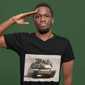 May include: A man wearing a black t-shirt with a printed image of a military tank in a desert setting. He is saluting with his right hand.