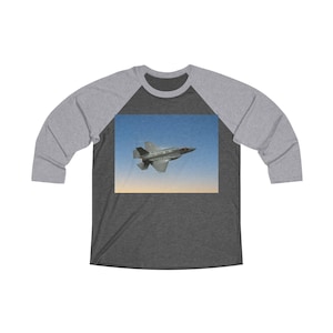 May include: A raglan baseball tee with a dark gray body and light gray sleeves. The front features a graphic of a gray jet fighter plane soaring in a blue sky with a yellow horizon. A casual, comfortable shirt.