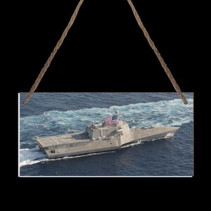 Puede incluir: Un letrero rectangular con un buque de guerra gris navegando sobre agua azul. El barco tiene una bandera estadounidense y crea estelas blancas y espumosas. El letrero está suspendido por dos cuerdas marrones.