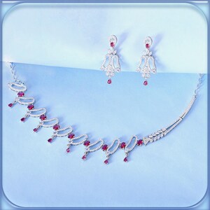 May include: A dazzling jewelry set featuring a necklace and matching earrings. The necklace has a delicate design with oval-shaped red gemstones and dangling teardrop-shaped gems. The earrings mirror the necklace's design, with red teardrop gems. The set is set against a light blue background.