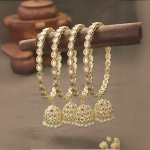 Könnte beinhalten: Vier goldfarbene Armreifen mit tropfenförmigen, klaren Steinen und glockenförmigen Anhängern. Die Anhänger sind mit aufwendigen Mustern und kleinen weißen Perlen verziert. Die Armreifen werden auf einem Holzstab präsentiert.