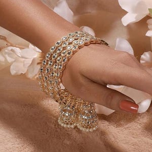 May include: A gold-toned bangle bracelet featuring a repeating teardrop pattern inlaid with clear stones. The bracelet includes two dangling bell-shaped charms at the base, each decorated with small clear stones and tiny white pearl-like beads.