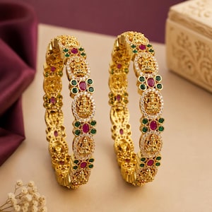 May include: A pair of gold-toned bangles featuring intricate embossed deity figures, alternating with circular clusters of small white stones and oval-shaped red and green stones. The metal surface is textured with fine detailing throughout the band.