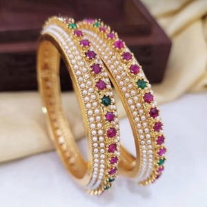 May include: Two gold bangles with intricate detailing. Each bangle is adorned with small, round white pearls and alternating ruby and emerald-colored gemstones. The bangles have a textured gold finish and are displayed against a soft, neutral background.