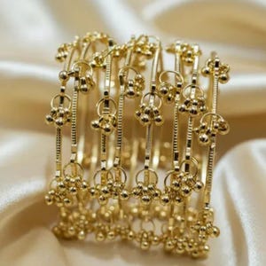 Puede incluir: Un conjunto de pulseras doradas. Cada pulsera presenta una serie de pequeñas cuentas redondas doradas y elementos decorativos. Las pulseras están dispuestas sobre una tela suave de color crema.