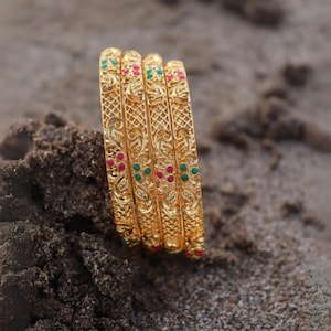 Peut inclure: Quatre bracelets dorés avec des motifs floraux et en treillis complexes. Chaque bracelet est orné de pierres précieuses rouges et vertes. Les bracelets sont présentés sur une surface sablonneuse, mettant en valeur leurs détails ornés et leurs couleurs vives.