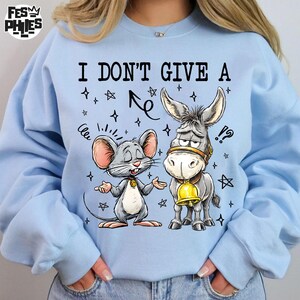 May include: Light blue sweatshirt with a graphic of a mouse and a donkey. The mouse has its arms out, and the donkey has a bell around its neck. The text "I DON'T GIVE A" is above the characters.