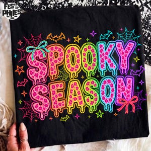 May include: A black t-shirt featuring the text SPOOKY SEASON in a dripping, multi-colored font with a pink floral pattern. The design is surrounded by neon-colored spider webs, stars, bats, and two bows in a vibrant, high-contrast graphic style.