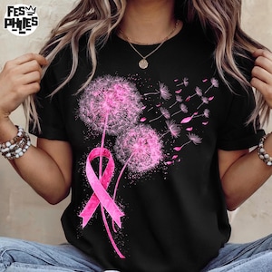 May include: Black t-shirt featuring a pink dandelion design with a pink ribbon. The dandelion has a fluffy, textured appearance, with some seeds floating away. The ribbon is a symbol of breast cancer awareness. The shirt has short sleeves.
