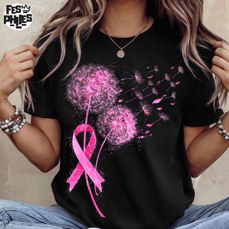 May include: Black t-shirt featuring a pink dandelion design with a pink ribbon. The dandelion has a fluffy, textured appearance, with some seeds floating away. The ribbon is a symbol of breast cancer awareness. The shirt has short sleeves.